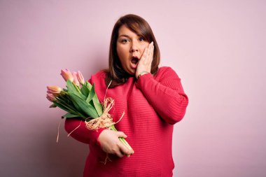 Güzel, iri yarı bir kadın pembe arka planda romantik laleler tutuyor. Korkmuş ve şok olmuş, şaşırmış ve şaşırmış bir halde.