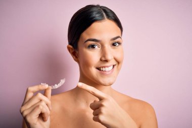 İzole edilmiş pembe arka plan üzerinde diş hizalayan genç ve güzel bir kız. Elle ve parmakla işaret ederek çok mutlu.
