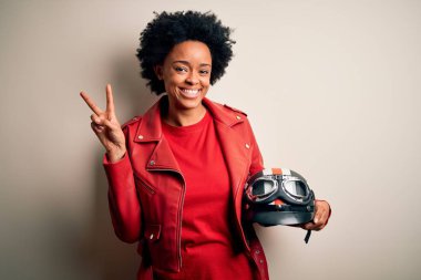 Kıvırcık saçlı, motosiklet kaskı takan, kameraya bakıp zafer işareti yapan Afro-Amerikan motosikletli genç bir kadın. İki numara..