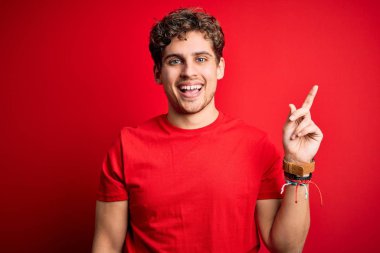 Sarışın, kıvırcık saçlı, kırmızı arka planda sıradan tişörtler giyen, yüzünde kocaman bir gülümseme olan, eli kameraya bakan genç ve yakışıklı bir adam..