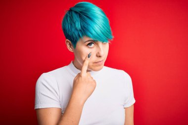 Mavi saçlı, kırmızı arka planda sıradan bir tişört giyen genç ve güzel bir kadın. Gözünü dikip jestini izliyor. Şüpheli bir ifade.