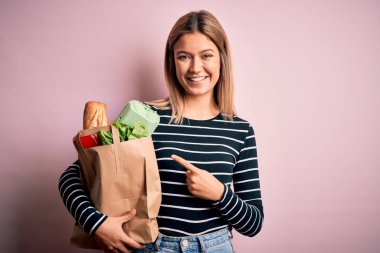 Genç ve güzel bir kadın, pembe arka plan üzerinden alışveriş torbası tutuyor. Elle ve parmakla işaret ederek çok mutlu.
