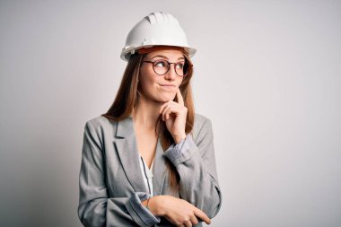 Beyaz arka plan üzerine güvenlik kaskı takan genç, güzel, kızıl saçlı bir mimar kadın. Elleri çenesinde soru sorarken, düşünceli bir ifade takınıyor. Düşünceli bir yüzle gülümsüyor. Şüphe kavramı.