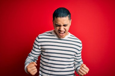 İzole edilmiş kırmızı arka planda dikilen sıradan çizgili tişört giyen genç Brezilyalı adam kolları havada, gülümseyen ve başarı için bağıran mutlu ve heyecanlı bir jest yapıyor. Kutlama konsepti.