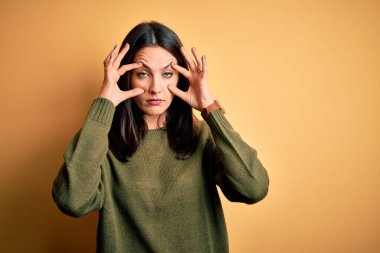 Sarı arka planda yeşil süveter giyen, mavi gözlü genç esmer kadın gözlerini parmaklarıyla açmaya çalışıyor, uykulu ve yorgun.