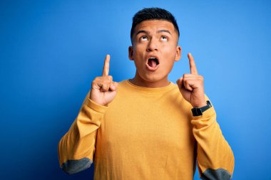 Sarı günlük süveter giyen genç, yakışıklı latin adam mavi arka planda şaşkın şaşkın yukarı bakıyor ve parmaklarıyla işaret ediyor ve kollarını kaldırıyordu..