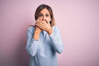 Orta yaşlı güzel bir kadın, günlük tişört giyip pembe arka planda dikiliyor. Ağzını yanlışlıkla ellerle kapatıyor. Gizli konsept.