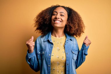 Afro saçlı, sarı arka plan üzerine günlük kot gömlek giyen genç Afrikalı Amerikalı kadın kollarını kaldırarak ve açık gözlerle başarıyı kutluyor ve şaşırıyor. Kazanan konsept.