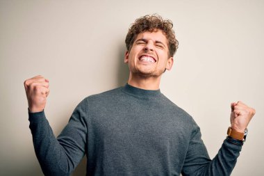 Sarışın, kıvırcık saçlı, beyaz arka planda rahat süveter giyen çok mutlu ve heyecanlı genç bir adam. Kolları havada, gülümseyen ve başarı için çığlık atan, kazanan bir jest yapıyor. Kutlama konsepti.