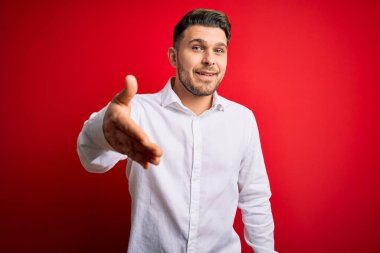 Mavi gözlü, zarif gömlekli genç bir iş adamı kırmızı izole edilmiş arka planda duruyor. Dostça bir selamlama ve hoş geldin hediyesi olarak gülümsüyor. Başarılı bir iş..