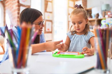 Beyaz kız çocuk, bayan öğretmenle oyun oynayıp öğreniyor. Anne ve kızı oyun odasında oyuncakların etrafında manyetik karatahtaya resim çiziyorlar.