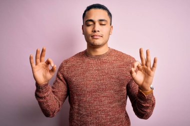 İzole edilmiş pembe arka plan üzerinde duran sıradan bir süveter giyen genç Brezilyalı adam rahatla ve parmaklarıyla meditasyon hareketi yaparken gözleri kapalı gülümse. Yoga konsepti.