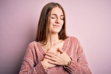Genç, güzel, kızıl saçlı bir kadın pembe arka planda rahat bir süveter giyiyor. Elleri göğsünde, gözleri kapalı ve yüzünde minnet dolu bir jest ile gülümsüyor. Sağlık kavramı.