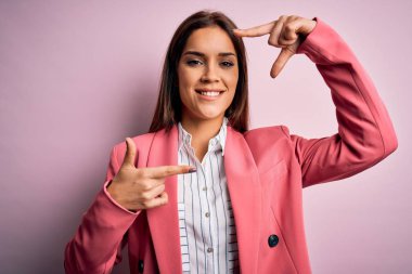 İzole edilmiş pembe arka planda duran ceketli güzel esmer kadın gülümsüyor ve mutlu yüzlü elleri ve parmakları olan bir çerçeve yapıyor. Yaratıcılık ve fotoğrafçılık kavramı.