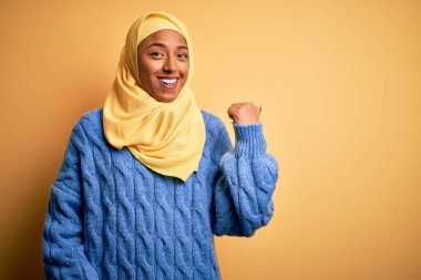 Genç Afrikalı Afro kadın, izole edilmiş sarı arka planda Müslüman başörtüsü takıyor. Gülümsüyor, gülümsüyor ve başparmağıyla yana bakıyor..