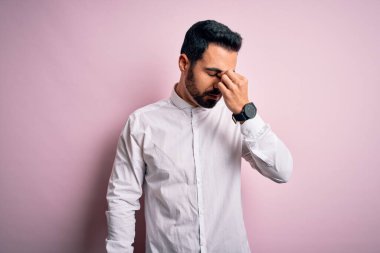 Sakallı, sıradan bir gömlek giyen, pembe arka planda duran, burnu ve gözleri ovuşturan, yorgun ve başı ağrıyan genç yakışıklı bir adam. Stres ve hüsran kavramı.