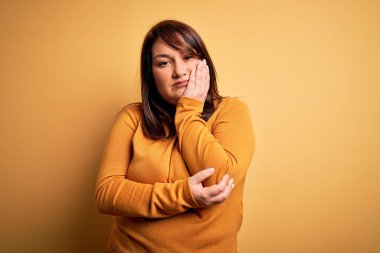 Güzel esmer ve cüsseli bir kadın izole edilmiş sarı arka plan üzerine günlük süveter giyip kollarını kavuşturarak depresyondan sıkılmış ve yorgun görünüyor..