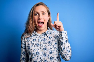 Mavi arka planda dikilmiş, gündelik tişört giyen genç ve güzel sarışın bir kadın başarılı bir fikri işaret ediyor. Heyecanlı ve mutlu. Bir numara..