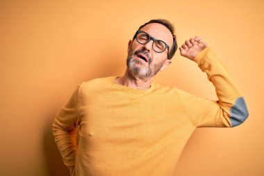 Orta yaşlı, ağarmış, günlük süveter ve gözlük takan, izole edilmiş sarı arka planda gerinen, yorgun ve rahat, uykulu ve sabahın erken saatleri için esneyen bir adam.