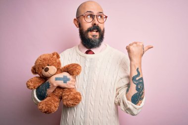 Sakallı ve dövmeli yakışıklı kel adam ayıcığı pembe arka planda tutuyor ve gülümseyen bir gülümsemeyle baş parmağıyla işaret ediyor.