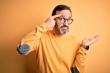 Orta yaşlı, beyaz bir adam, sarı arka planda günlük süveter ve gözlük takıyor. Avuç içinin açık olması, fotokopi alanını göstermesi ve alnını işaret etmesi kafasını karıştırıyor ve rahatsız ediyor. Bunu bir düşün..