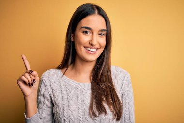 Sarı arka plan üzerine beyaz süveter giyen genç ve güzel esmer kadın bir numaralı parmağını gösterirken kendinden emin ve mutlu bir şekilde gülümsüyor..