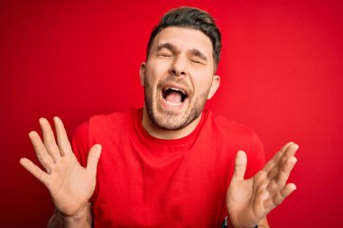 Mavi gözlü genç adam kırmızı izole edilmiş arka plan üzerine gündelik tişört giyerek çılgın ve çılgın başarısını kollarını kaldırarak ve gözlerini kapayarak heyecanla bağırarak kutluyor. Kazanan konsept