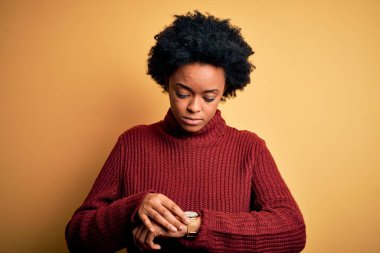 Kıvırcık saçlı, balıkçı yaka kazak giyen genç, güzel Afro-Amerikan kadın. Kol saatini kontrol ediyor, rahat ve kendinden emin.