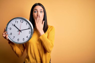 Mavi gözlü genç kadın geri sayım yapıyor sarı arka plan üzerinde büyük bir saat tutuyor ağzı kapalı hata üstüne utanç, korku ifadesi, sessizlik içinde korkmak, gizli bir konsept.