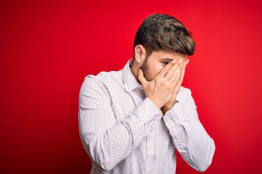 Sarışın, sakallı ve mavi gözlü genç bir işadamı kırmızı arka planda zarif bir gömlek giyiyor ve ağlarken suratını ellerle kapatıyor. Depresyon kavramı.