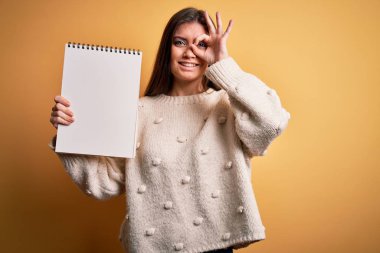 Mavi gözlü genç bir kadın sarı arka planda boş beyaz bir defter tutuyor. Gülümsüyor. Tamam işaret veriyor.