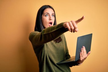 Mavi gözlü genç esmer kadın sarı arkaplanda bilgisayar bilgisayarı kullanarak çalışıyor. Parmağıyla işaret ediyor. İleride şaşırmış, ağzı açık, ön tarafta bir şey var.