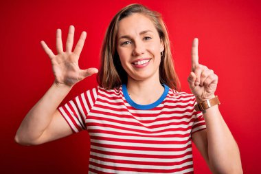 Genç ve güzel sarışın bir kadın, izole edilmiş kırmızı arka plan üzerinde gündelik çizgili tişört giyiyor ve 6 numaralı parmaklarıyla işaret ederken kendinden emin ve mutlu gülümsüyor..