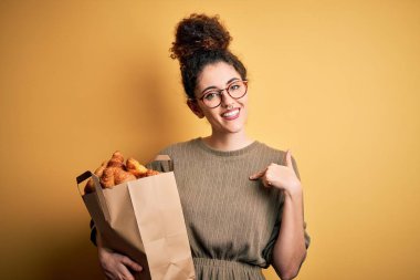 Kıvırcık saçlı ve elinde kruvasan dolu kruvasanlarla elinde piercing olan güzel bir kadın kendini işaret ediyor.