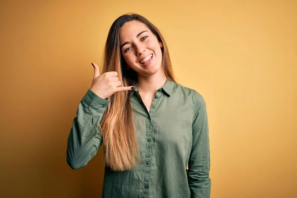 Young beautiful blonde woman with blue eyes wearing green shirt over yellow background smiling doing phone gesture with hand and fingers like talking on the telephone. Communicating concepts.