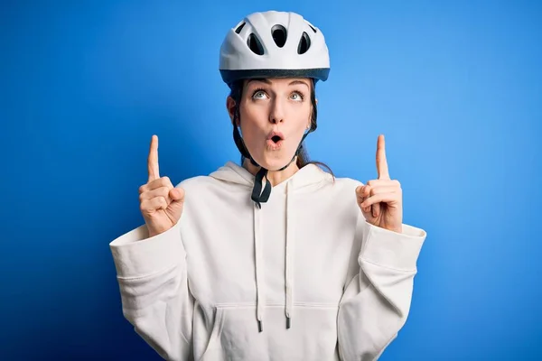 Young beautiful redhead cyclist woman wearing bike helmet over isolated blue background amazed and surprised looking up and pointing with fingers and raised arms.
