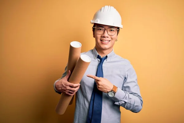 Young handsome chinese architect man wearing glasses and safety helmet holding blueprints very happy pointing with hand and finger