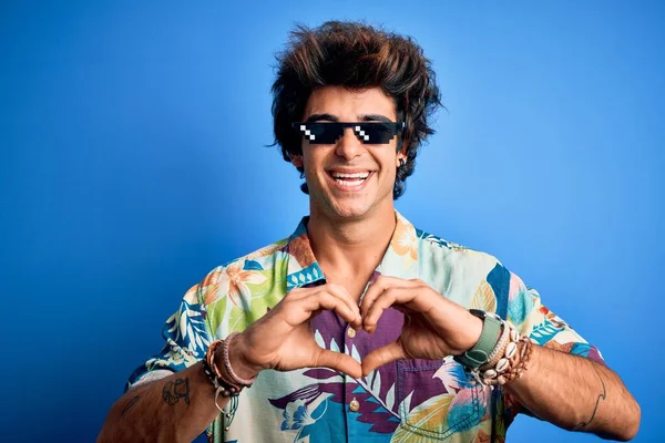 Young handsome man wearing thug life glasses standing over isolated blue background smiling in love showing heart symbol and shape with hands. Romantic concept.