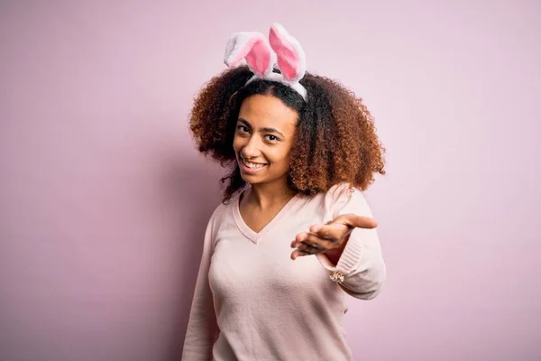 ピンクの背景の上にウサギの耳を身に着けているアフロの髪を持つ若いアフリカ系アメリカ人女性は 挨拶や歓迎として握手を提供しています 成功事業 — ストック写真