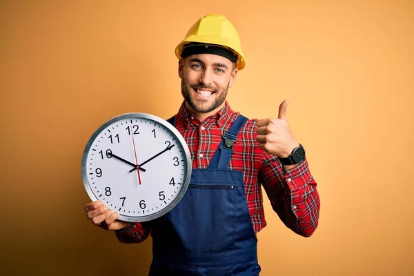 Junger Bauarbeiter Mit Schutzhelm Hält Große Uhr Über Gelbem Hintergrund — Stockfoto