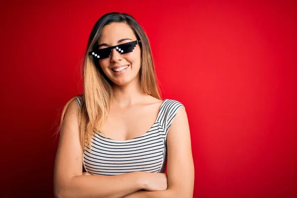 Young beautiful brunette woman wearing funny thug life sunglasses over red background happy face smiling with crossed arms looking at the camera. Positive person.