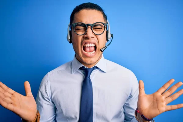 Young brazilian call center agent man wearing glasses and tie working using headset celebrating mad and crazy for success with arms raised and closed eyes screaming excited. Winner concept