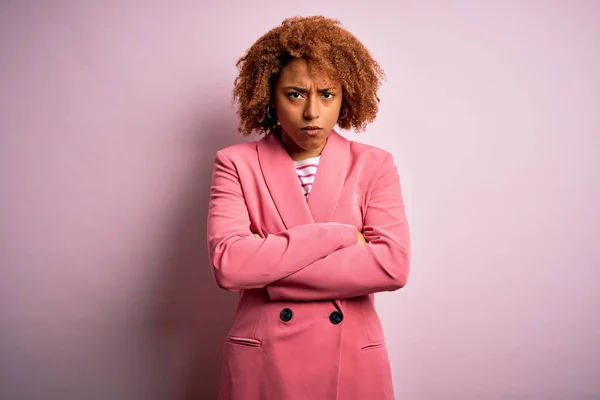 Young beautiful African American businesswoman with curly hair wearing elegant pink jacket skeptic and nervous, disapproving expression on face with crossed arms. Negative person.