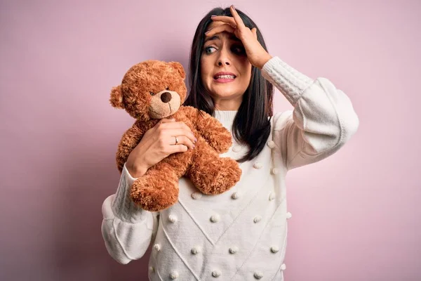 Young brunette woman with blue eyes hugging teddy bear stuffed animal over pink background stressed with hand on head, shocked with shame and surprise face, angry and frustrated. Fear and upset for mistake.