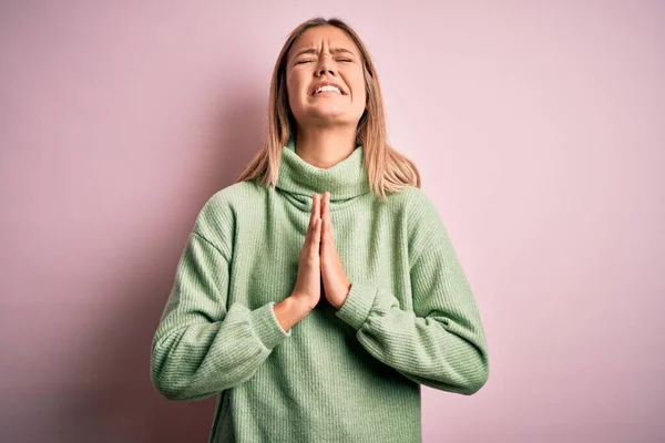 Young beautiful blonde woman wearing winter wool sweater over pink isolated background begging and praying with hands together with hope expression on face very emotional and worried. Begging.