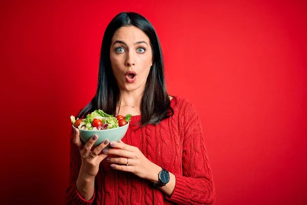 若いですブルネット女性ともに青い目食べる健康的な緑のサラダ上隔離された赤の背景怖がってショックで驚きの顔 恐怖と興奮で恐怖の表現 — ストック写真