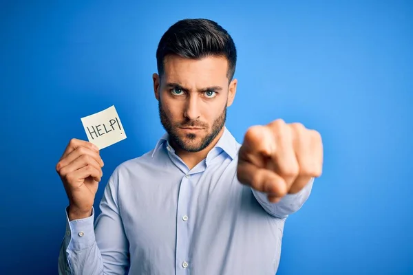 Young overworked business man asking for help holding paper over blue background pointing with finger to the camera and to you, hand sign, positive and confident gesture from the front