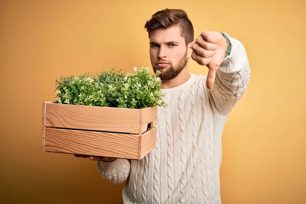 Young blond gardener man with beard and blue eyes holding wooden box with plants with angry face, negative sign showing dislike with thumbs down, rejection concept