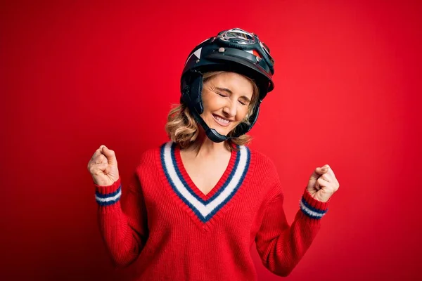 中世の美しいブロンドのオートバイの女性は 赤い背景の上に元ヘルメットを身に着けています非常に幸せと腕を上げて勝者のジェスチャーを行う興奮 笑顔と成功のために叫んで お祝いのコンセプト — ストック写真