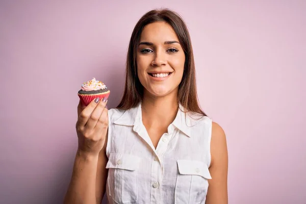 若いです美しいですブルネット女性食べるチョコレートカップケーキ上の隔離されたピンクの背景で幸せな顔立ちと笑顔で自信を持って笑顔示す歯 — ストック写真